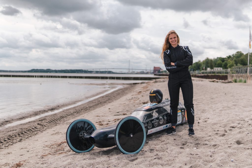 Image Kampagne für den Studiengang Regenerative Energien an der Hochschule Stralsund. Das Bild zeigt eine junge Studentin neben einem Wasserstoff-basierten Racing Car am Strand